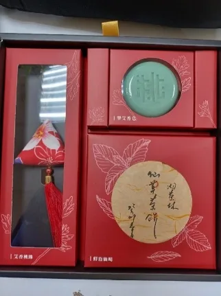 打開的紅色禮盒俯拍，左側為布製香包吊飾，右上為圓形艾草皂，右下為手寫題字的圓形仙草茶餅