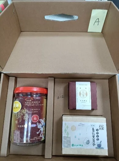 打開的牛皮紙禮盒俯拍，盒蓋貼有黃色標籤「A」，盒內放著一罐醬料、一盒金元寶造型香皂與一盒立體茶包