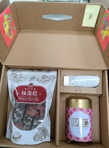 打開的牛皮紙禮盒俯拍，盒蓋貼有黃色標籤「B」，盒內放著一包核棗糕、一支乾洗手凝膠與一罐粉紅色茶葉罐