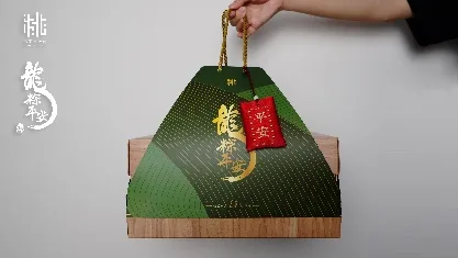 一隻手提著深綠色粽形禮盒，盒面印有金色「龍粽平安」字樣，繫著紅色平安符吊牌