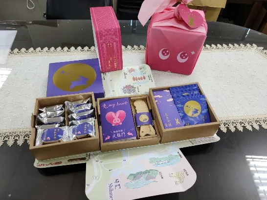桌面上攤開的中秋禮盒，含兩盒包裝糕點、一盒紫色月餅、一張石門水庫導覽摺頁，後方立著粉紅色布巾包裹的禮盒