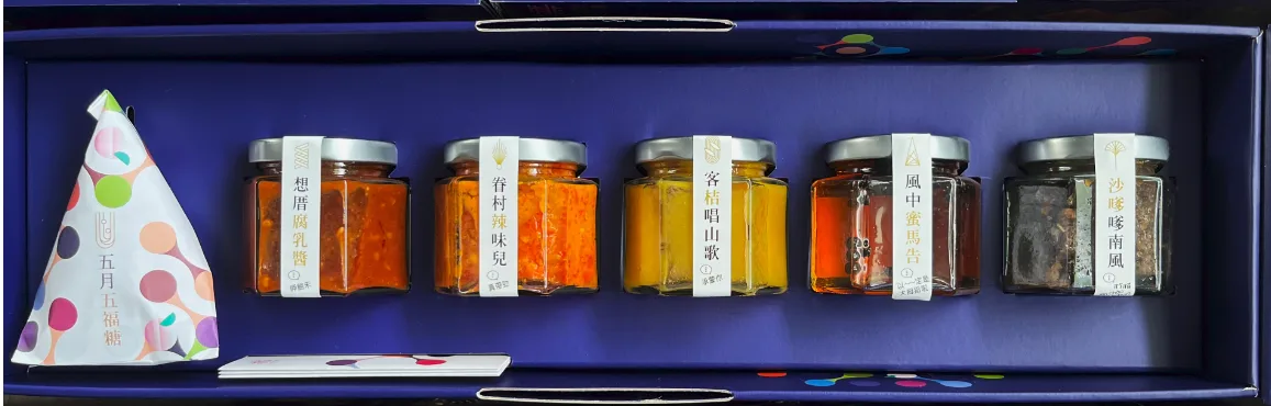 打開的深藍色禮盒俯拍，盒內排列五罐貼著手寫風標籤的玻璃醬料罐，左側放著彩色圓點包裝的粽形吊飾